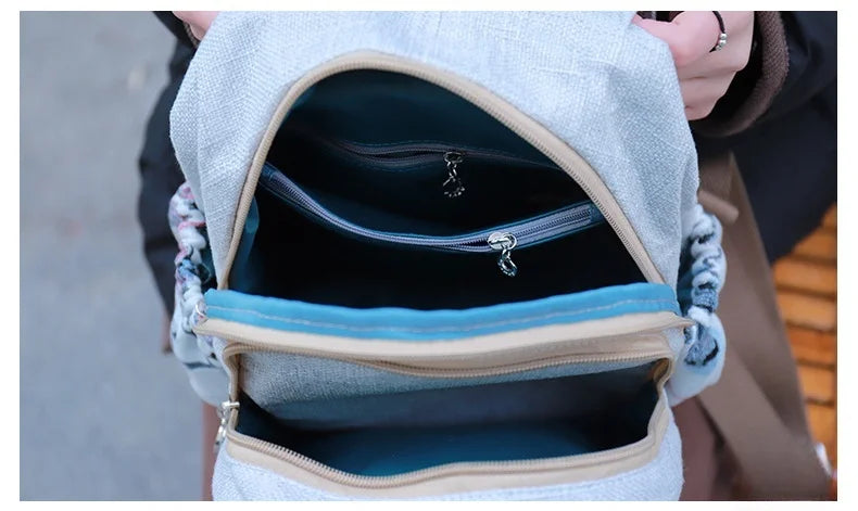 A light gray linen backpack is shown open, revealing a teal lining and decorative silver embroidery.