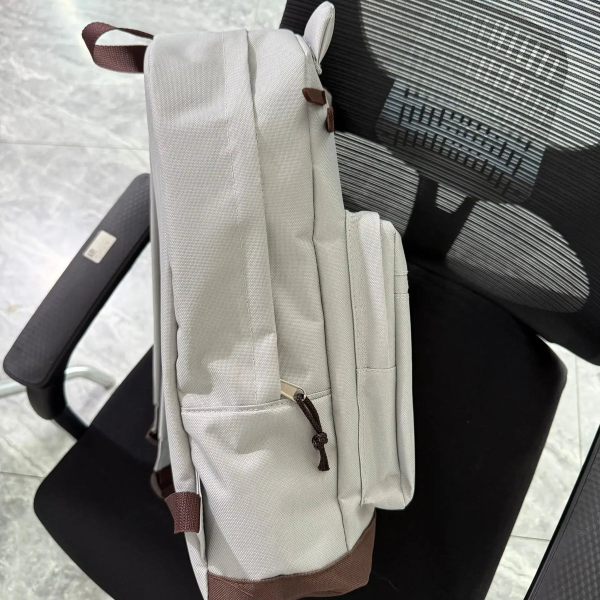 A light beige canvas backpack with brown accents resting on a black office chair.