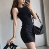 A woman wearing a black sleeveless bodycon tank dress is posing indoors.