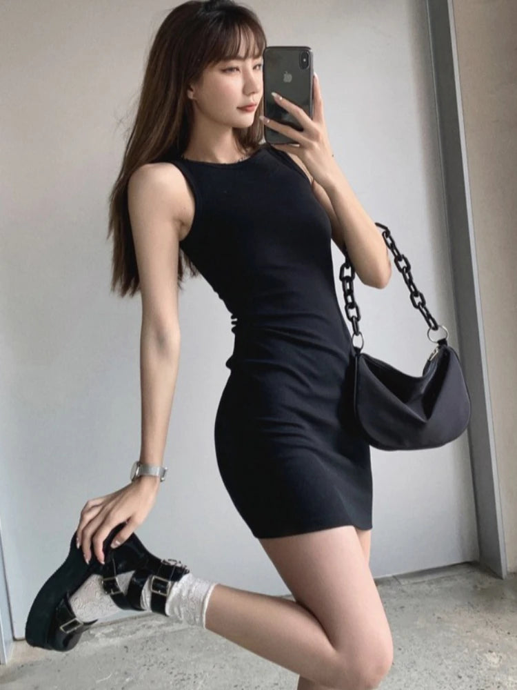 A woman wearing a black sleeveless bodycon tank dress is posing indoors.