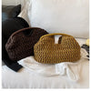 A close-up of a brown straw woven tote handbag resting on white fabric and bedding.