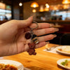 A hand holds a brown bear pendant keychain with a dark bead and a small coffee-themed charm.