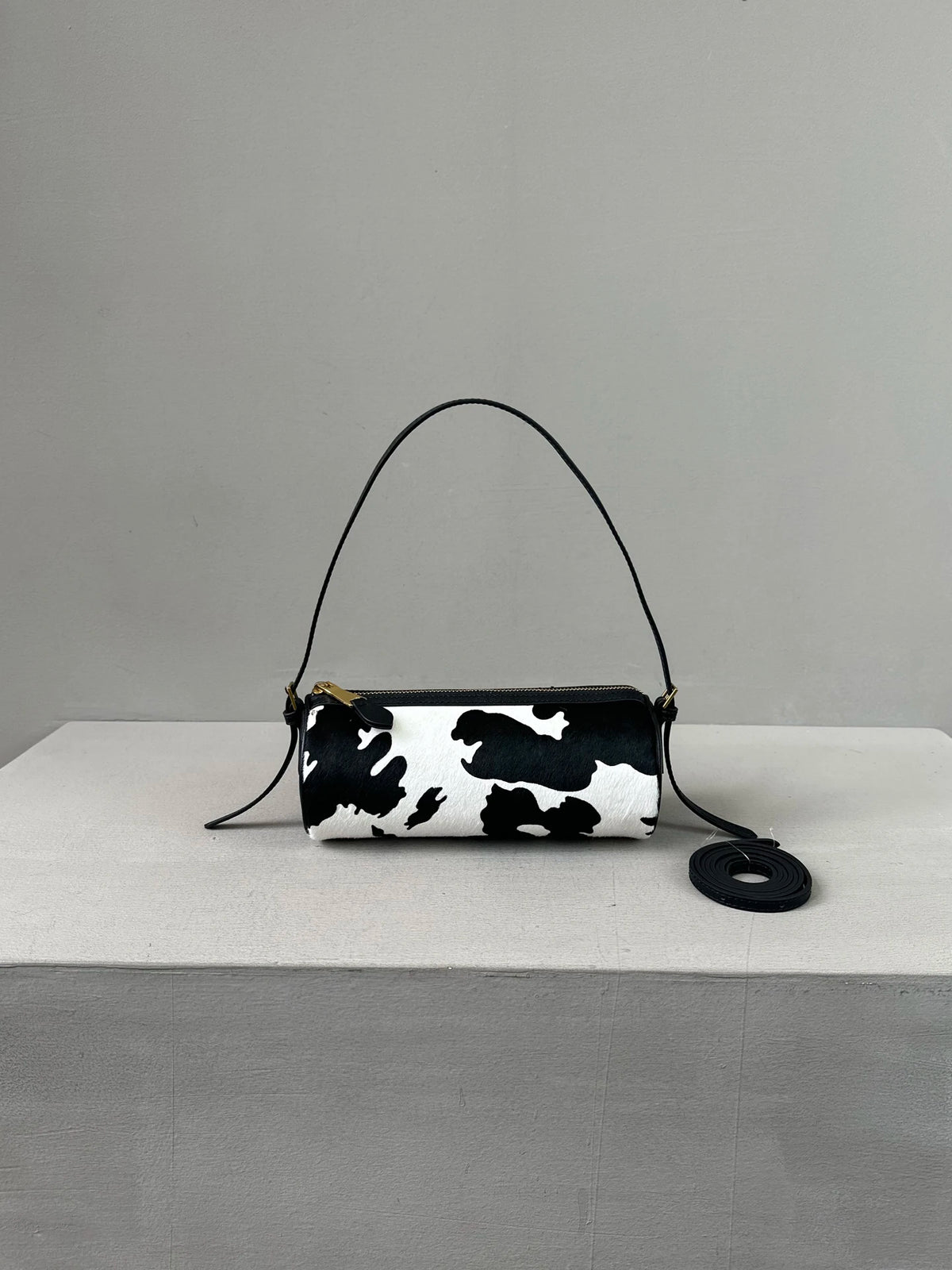 A black and white cow print cylinder shoulder bag with a thin black strap resting on a light gray surface.