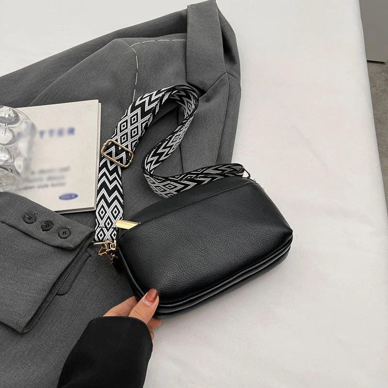 A woman's hand is holding a black vegan PU crossbody bag with a patterned gold chain strap, next to a gray blazer.