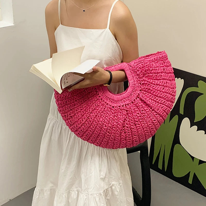 A woman holds a bright pink woven rattan half-moon beach handbag and an open book.