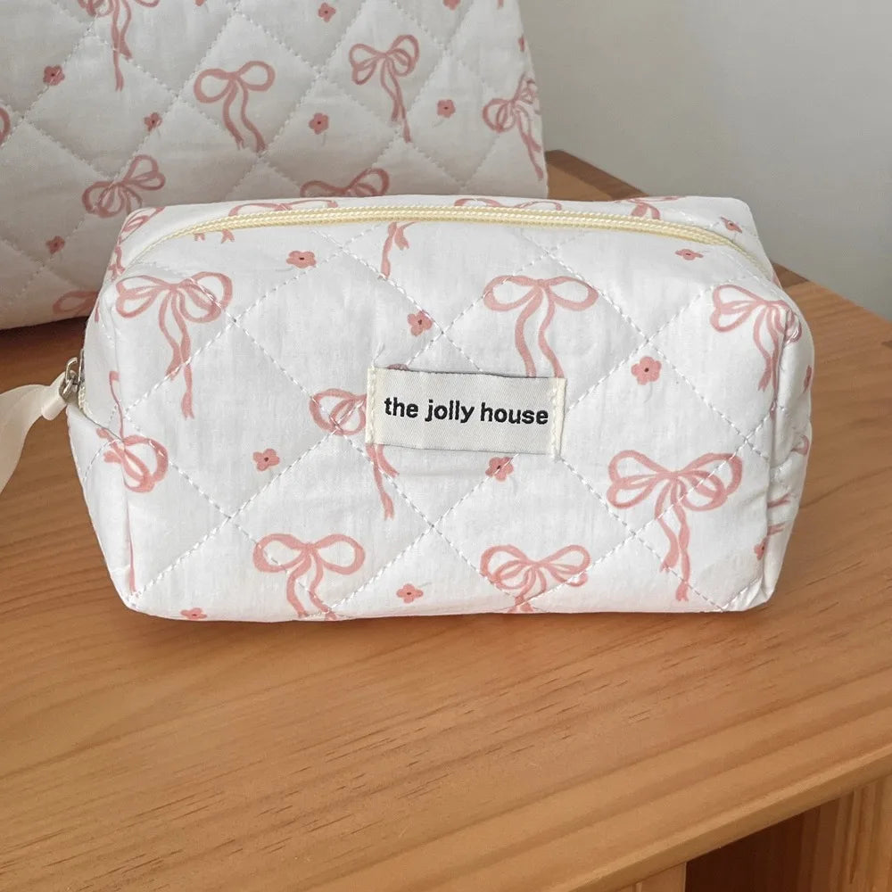 A white quilted cosmetic bag with pink ribbon embroidery rests on a wooden surface.