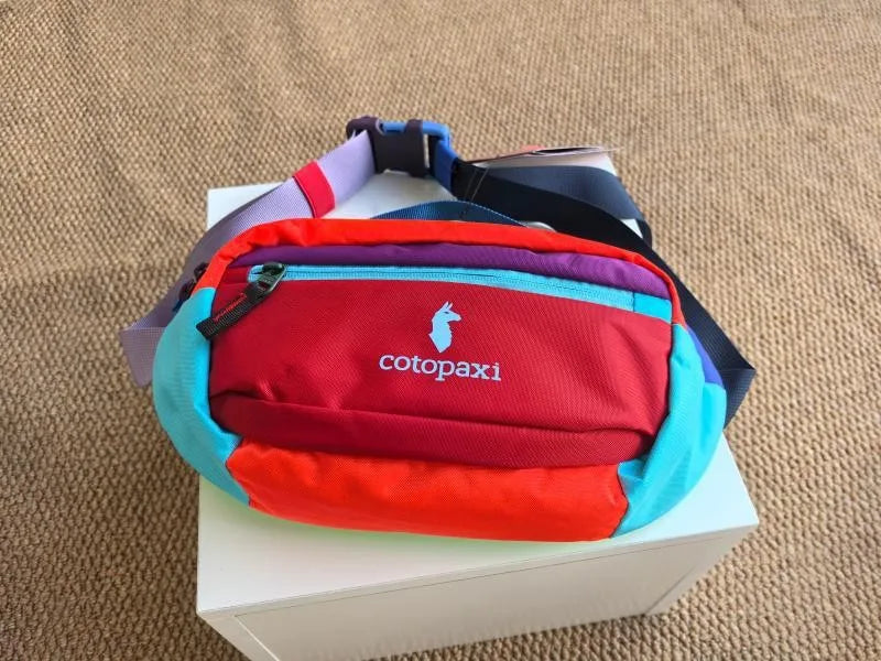 Rote und blaue Crossbody-Tasche mit lila Träger auf einer weißen Box.