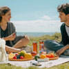 A young woman and man sitting on a blanket outdoors sharing fresh fruit and food with an insulated lunch bag.