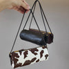 A brown and white cow print cylinder shoulder bag is being held by a hand against a gray background.