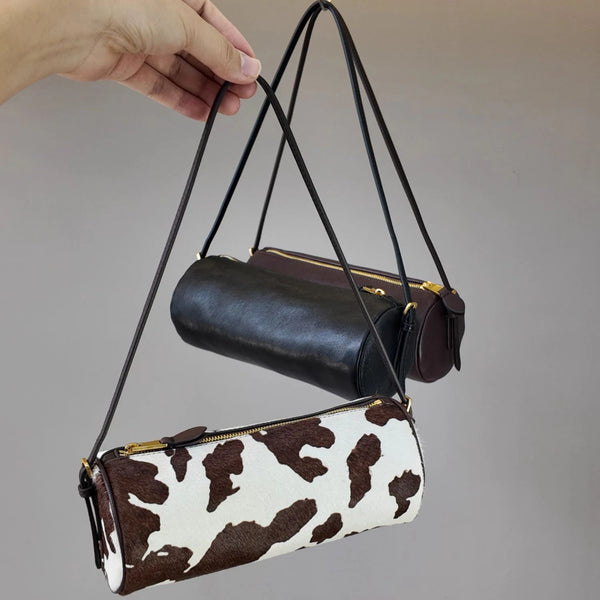 A brown and white cow print cylinder shoulder bag is being held by a hand against a gray background.