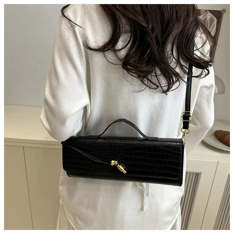 A black crocodile-embossed clutch bag is held by a person wearing a white top.