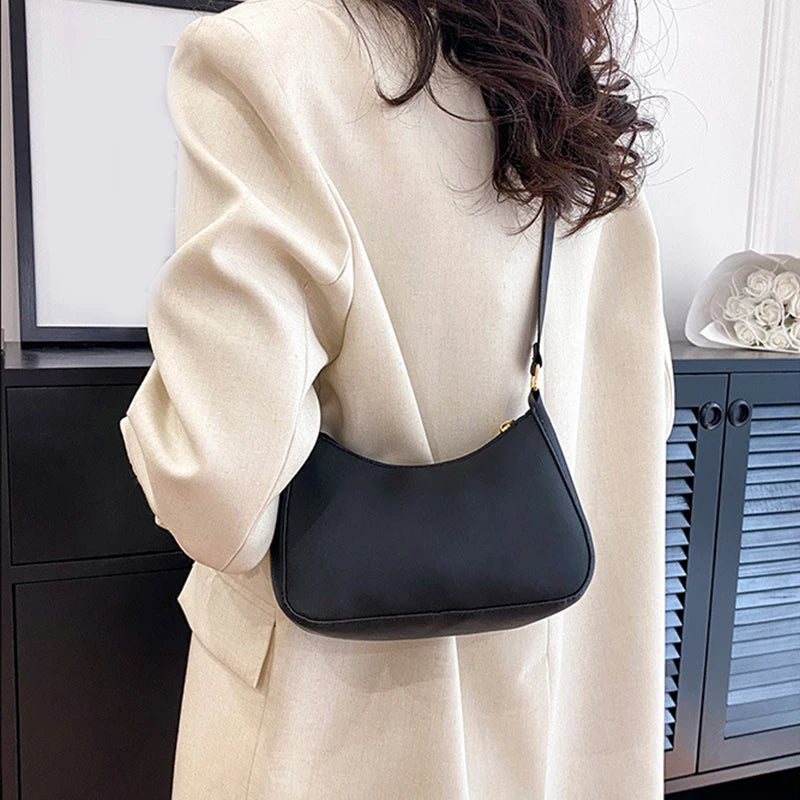 A beige coat sleeve shows a black leather shoulder bag with a gold strap against a dark cabinet.
