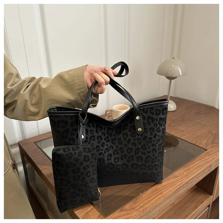A black leopard print tote bag with a leather handle is being held by a person on a wooden surface.