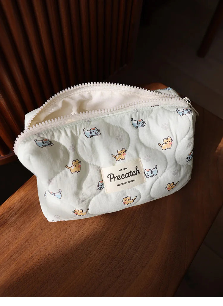 A white quilted cosmetic storage pouch with small printed cats resting on a wooden surface.