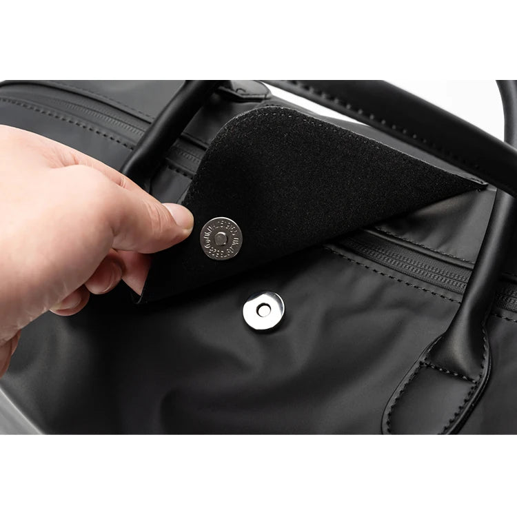 A hand is opening a flap on a black durable nylon travel duffle bag.