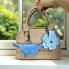 A beige PU leather handbag with a blue floral charm and keychain, held by a hand in front of a window.