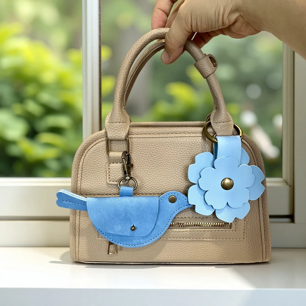A beige PU leather handbag with a blue floral charm and keychain, held by a hand in front of a window.