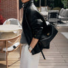 A woman wearing a black leather square shoulder crossbody bag and white pants standing outdoors on a wooden deck.