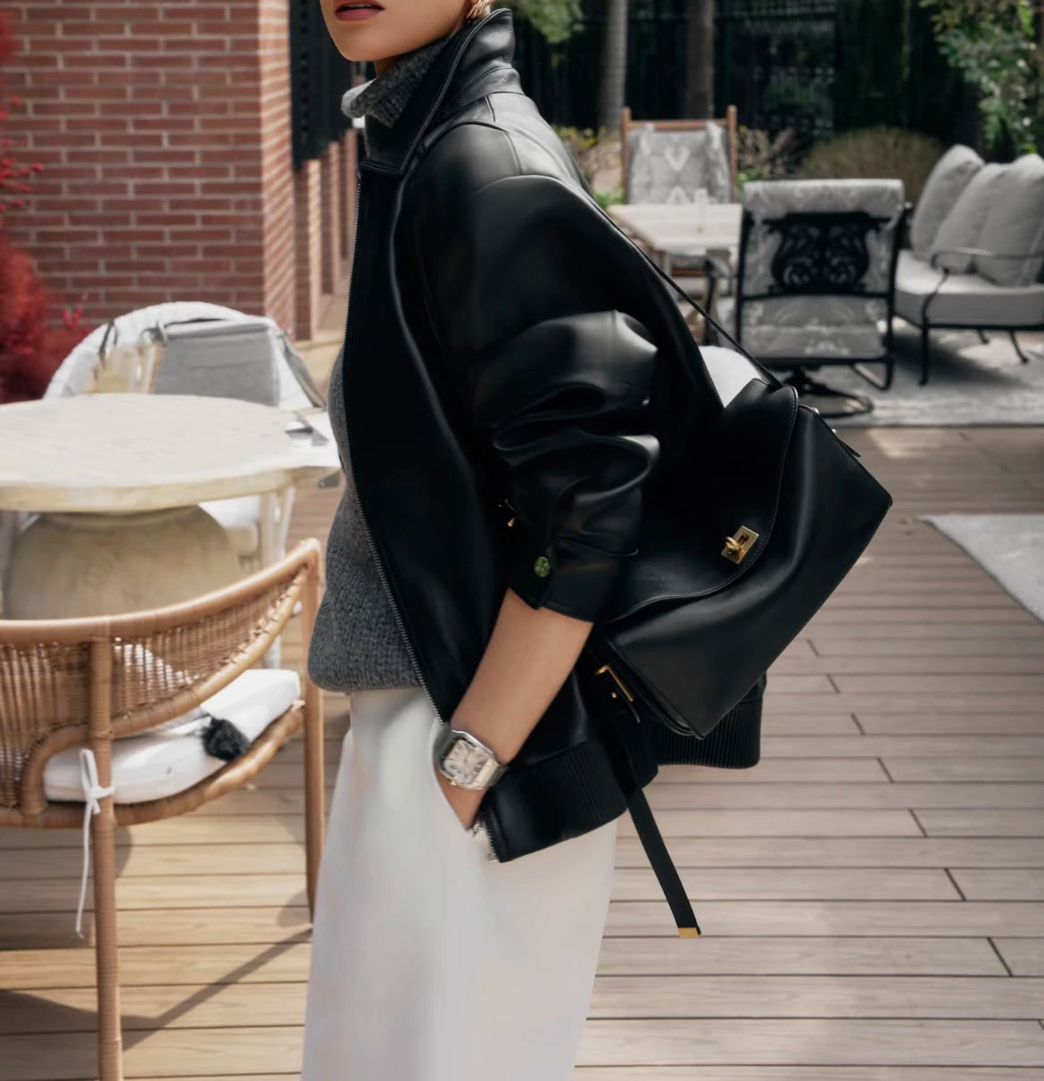 A woman wearing a black leather square shoulder crossbody bag and white pants standing outdoors on a wooden deck.