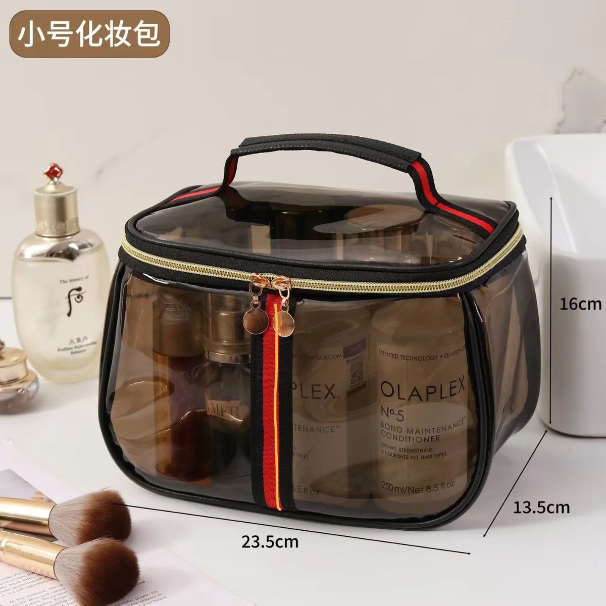 A clear waterproof travel toiletry bag with a black lid and handle, next to a bottle of liquid and some makeup brushes.