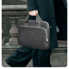 Une personne en costume noir tient une housse de laptop gris foncé et premium.
