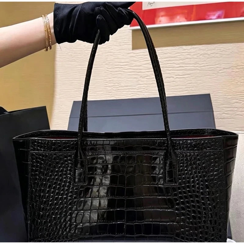 A black crocodile embossed leather tote bag being held by a hand wearing a black glove.
