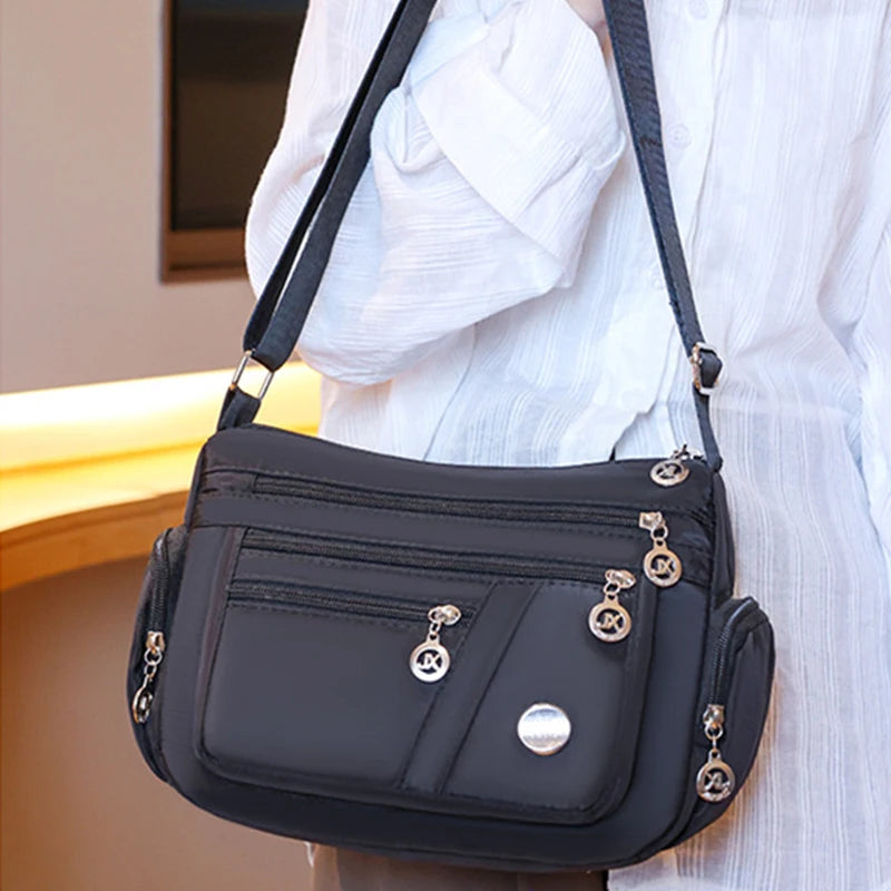 A black waterproof nylon crossbody tote bag with gold hardware is shown against a white shirt.