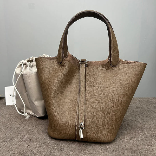A tan genuine leather bucket bag with a handle is displayed on a gray surface next to a light fabric pouch.
