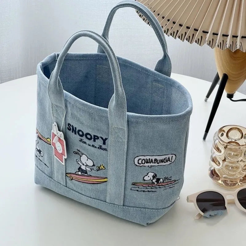 A light blue canvas shoulder bag featuring Snoopy cartoon designs and a straw sun hat in the background.