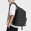 A young man wearing a beige shirt and black pants is holding a black Oxford school backpack with a laptop sleeve.
