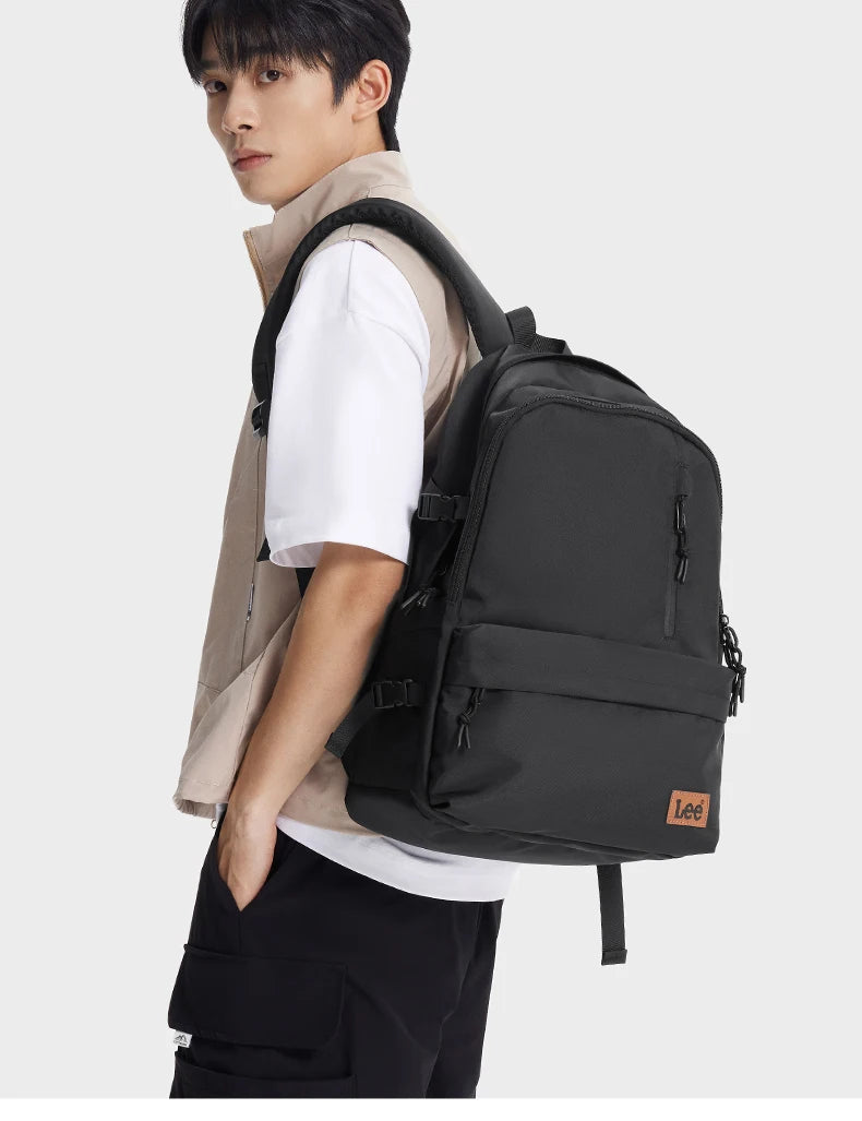 A young man wearing a beige shirt and black pants is holding a black Oxford school backpack with a laptop sleeve.