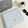 A light blue quilted laptop sleeve is shown next to a silver laptop and a glass perfume bottle on a white surface.