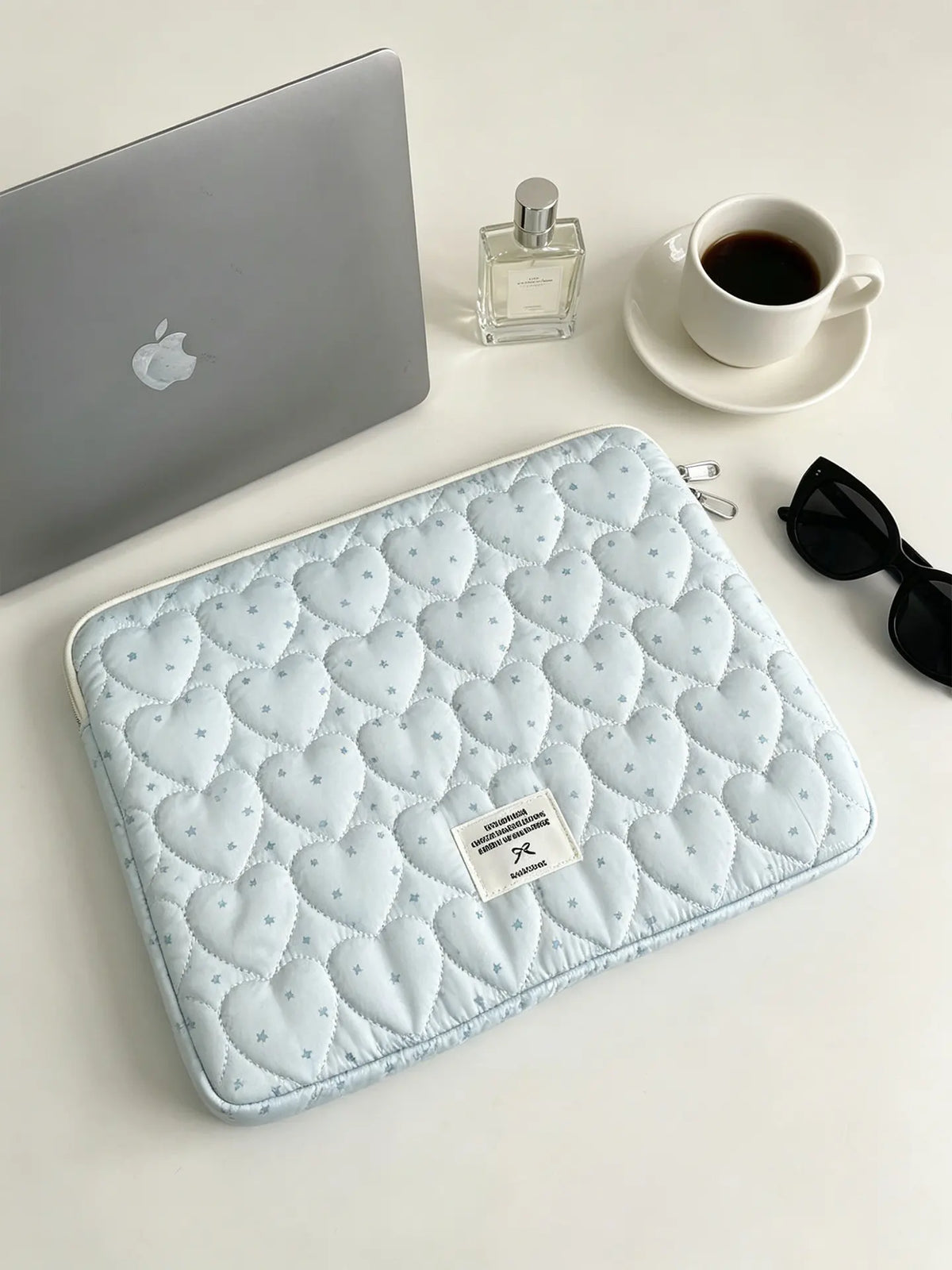 A light blue quilted laptop sleeve is shown next to a silver laptop and a glass perfume bottle on a white surface.