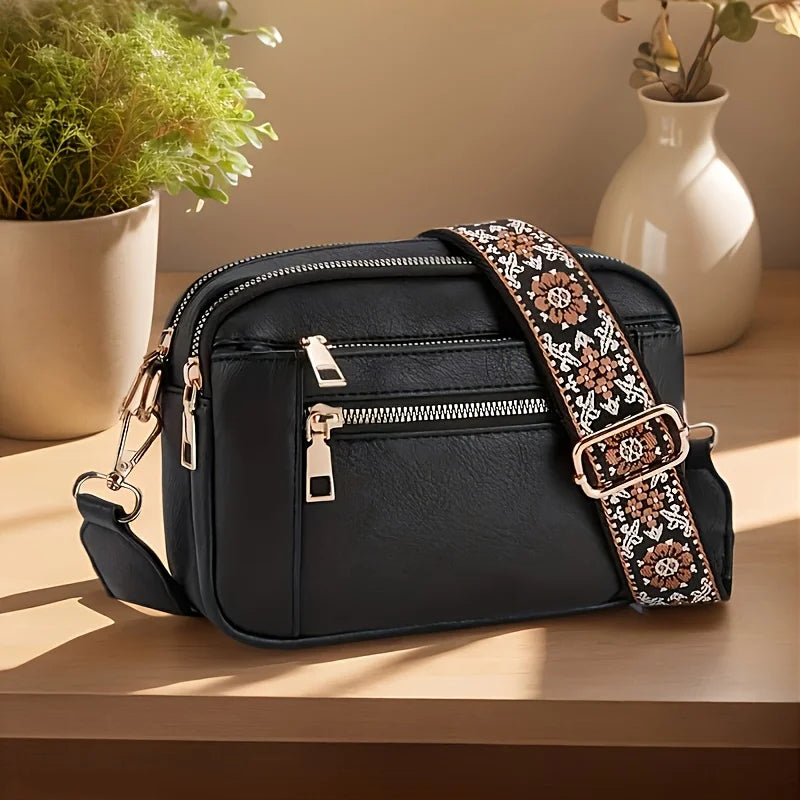 A black PU leather crossbody bag with a patterned strap rests on a wooden surface next to a potted plant.