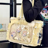 A light yellow transparent shoulder tote bag with decorative elements and a fluffy pompom, held by a person.