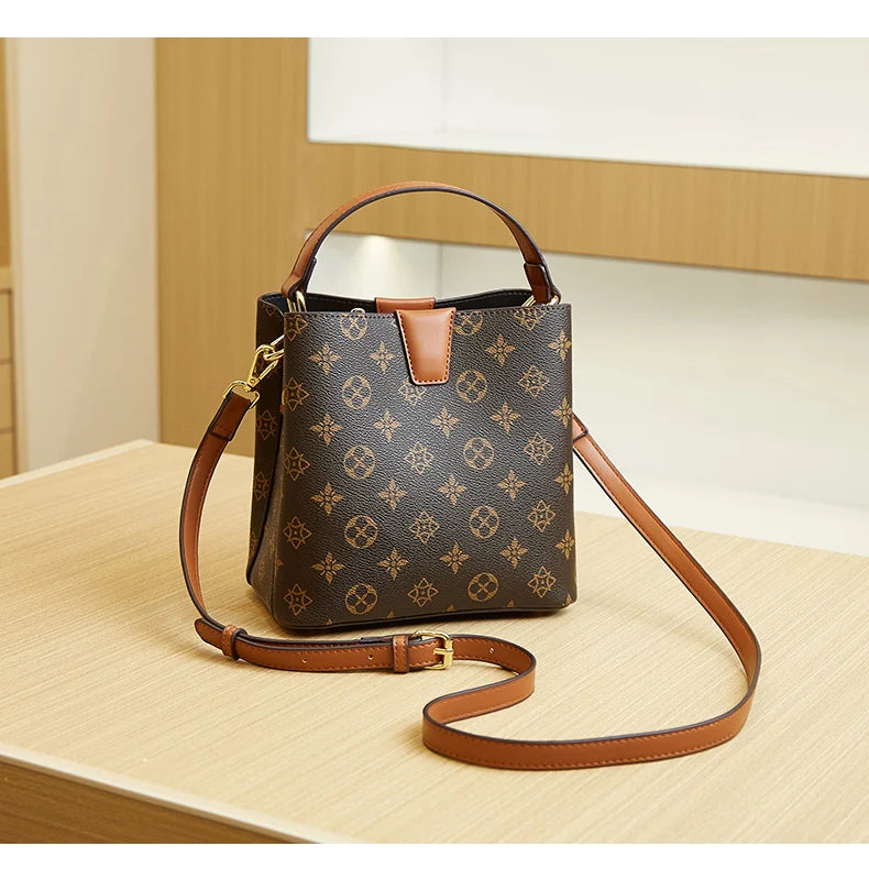 A brown leather bucket bag with gold hardware and a tan strap resting on a light wooden surface.
