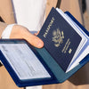 A hand holds a navy blue passport holder wallet with a gold embossed United States passport inside.