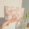 A hand holds a floral canvas makeup bag with pink and green embroidery against a white background.