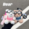 A white plush bear with pink accents is shown next to a brown plush bear, both featuring camera and glasses keychains.