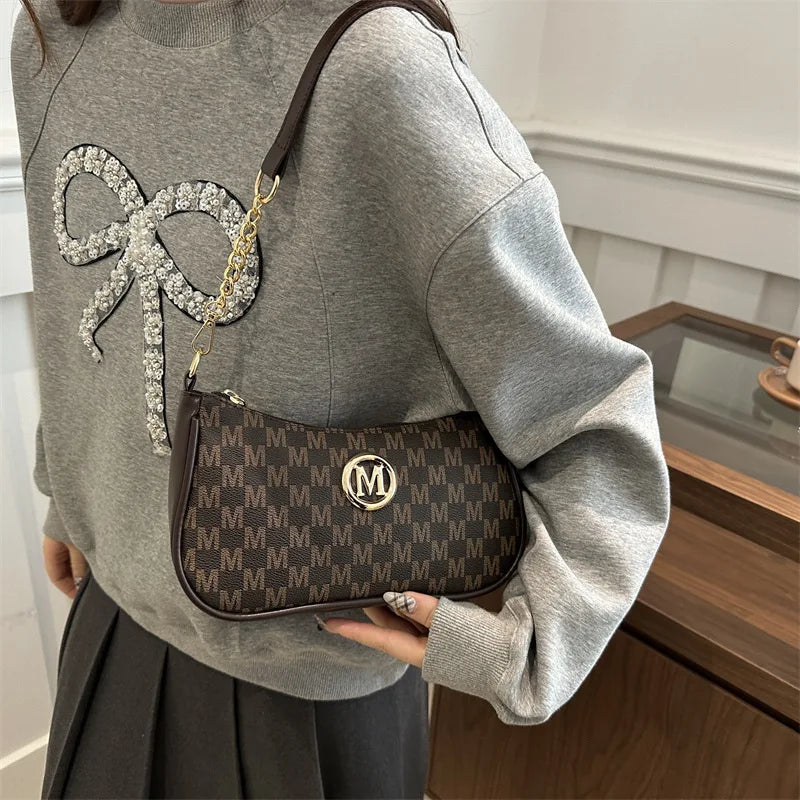 Una manga de sudadera gris muestra un bolso cruz de cuero marrón con una cadena dorada y un gran dije de M plateado.