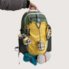A yellow and green durable laptop backpack with various tools and gear attached, being held by a hand.
