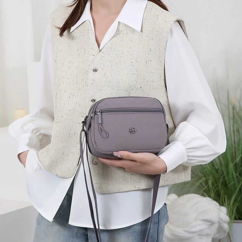 A woman wearing a cream vest and white shirt holds a mauve nylon crossbody bag.