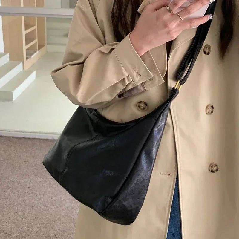 A hand holds a black vintage PU leather tote bag with a black crossbody strap against a light beige coat.