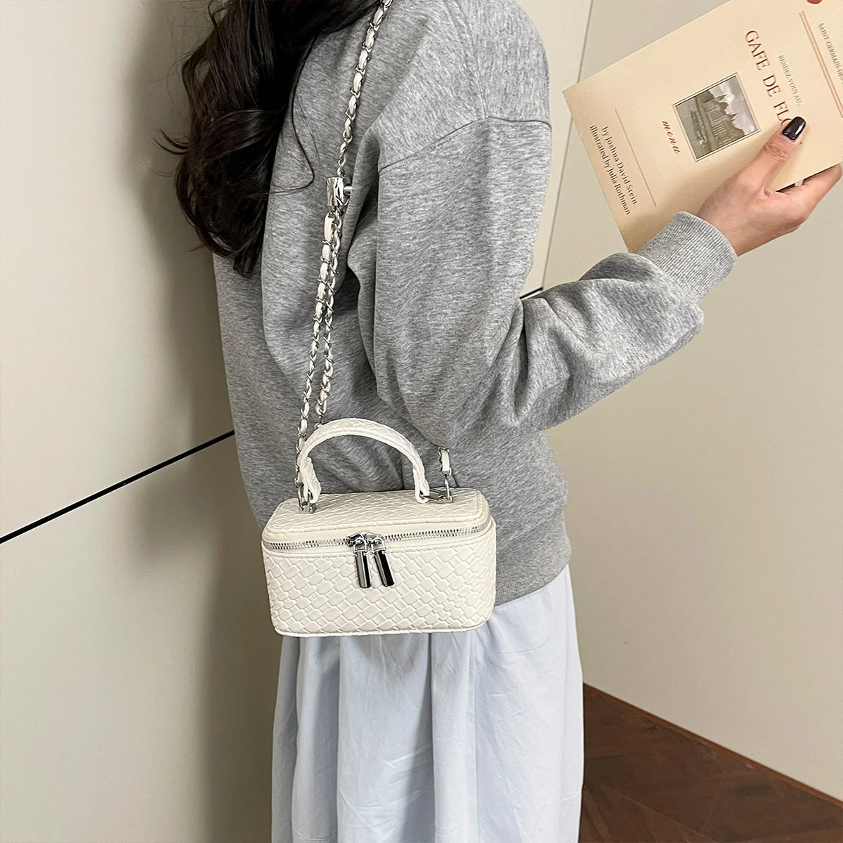 A person holds a cream woven crossbody bag with an adjustable silver chain strap and a light gray sweatshirt.