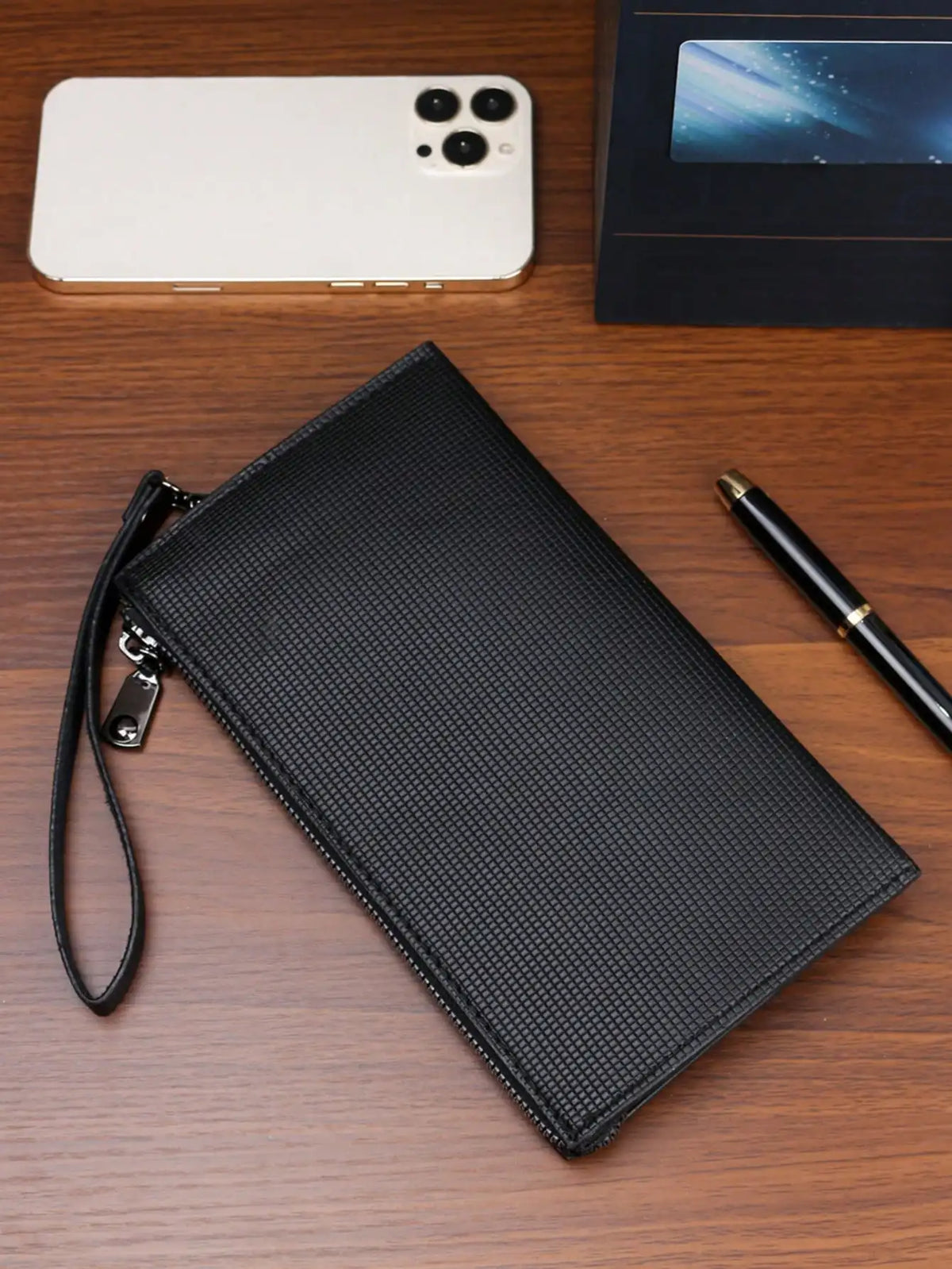 A black clutch bag with a textured surface is placed on a wooden table next to an iPhone.