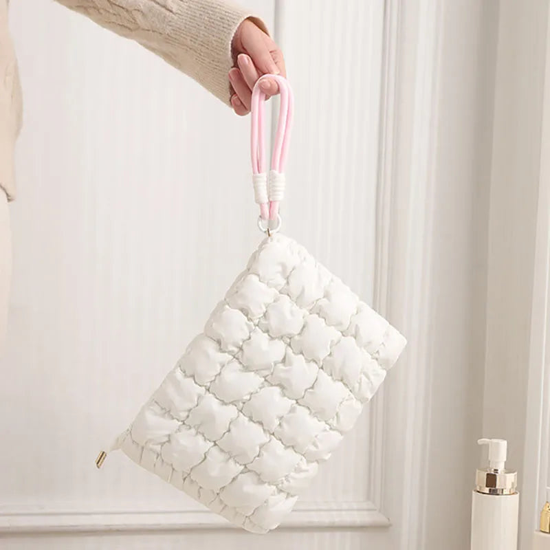 A white quilted puffer makeup bag with pink strap is being held by a hand.