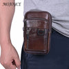 A brown vintage cowhide leather crossbody bag is shown being held by a person against a gray shirt and dark pants.