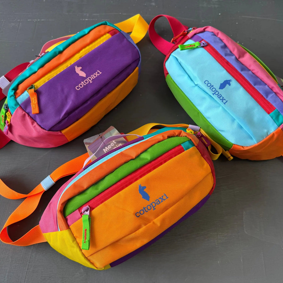 Drei farbenfrohe Cotopaxi Outdoor-Sport-Crossbodytaschen in lila, orange und blau.