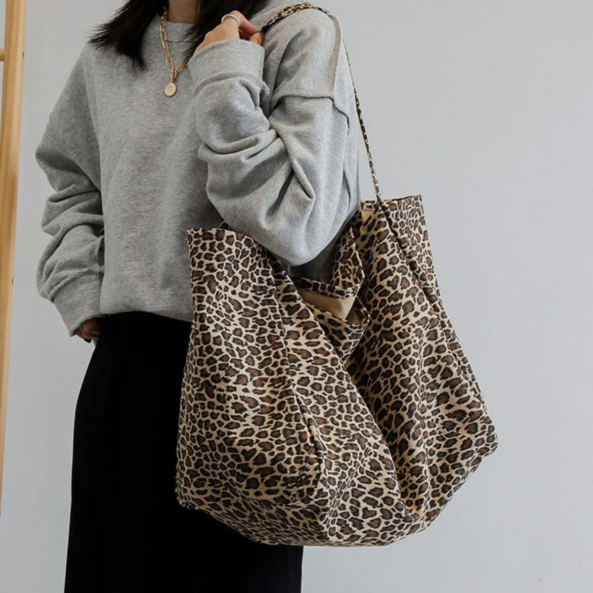 A person wearing a gray sweatshirt and black pants holds a leopard print canvas crossbody tote bag.