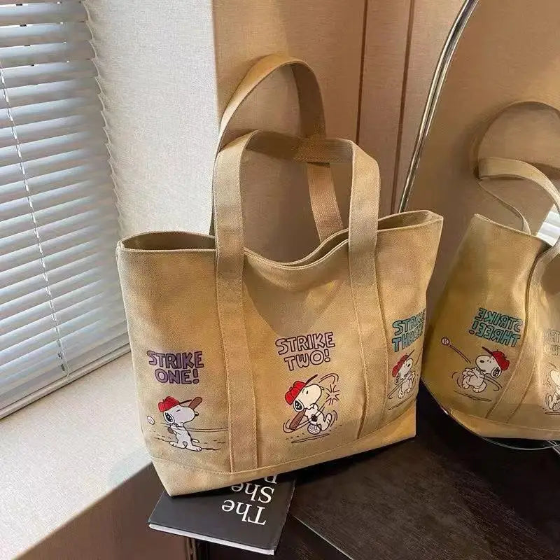 A tan canvas shoulder bag featuring Snoopy cartoon designs, displayed next to a window blind.